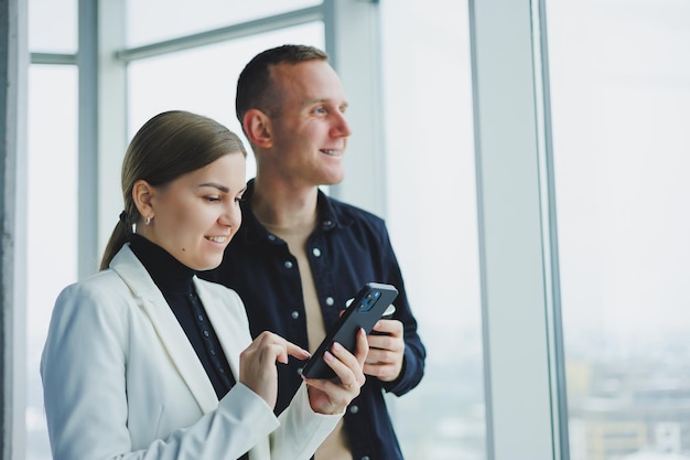 Homme et femme souriants debout près de la fenêtre avec des gadgets dans un bureau transparent moderne Conversation de collègues de travail dans un cadre informel