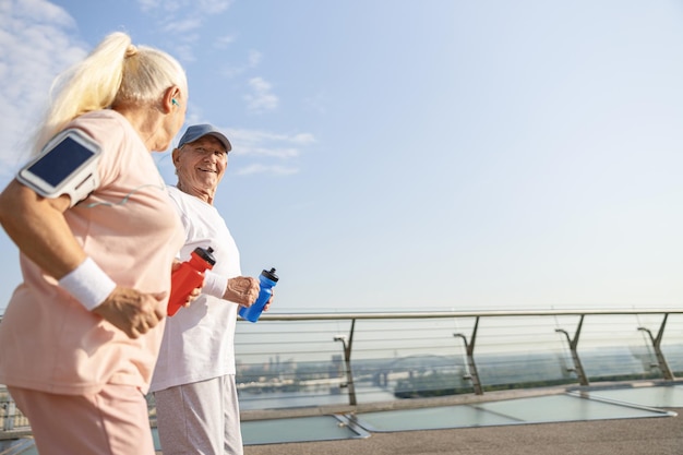 Un homme et une femme supérieurs joyeux avec des bouteilles d'eau longent la passerelle de la ville