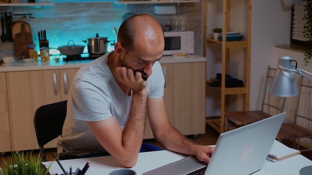 Homme fermant les yeux tout en travaillant de la cuisine à domicile pendant une date limite tard dans la nuit. Employé distant épuisé et occupé faisant la sieste sur une chaise se réveillant en travaillant sur un ordinateur portable à l'aide de la technologie moderne faisant des heures supplémentaires.