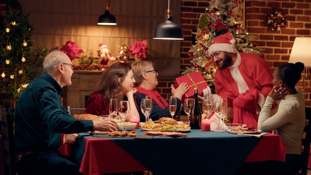 Homme festif déguisé en Père Noël donnant des cadeaux aux membres de la famille tout en profitant du dîner de Noël. Personne portant un costume traditionnel offrant des cadeaux aux gens tout en célébrant les vacances d'hiver.