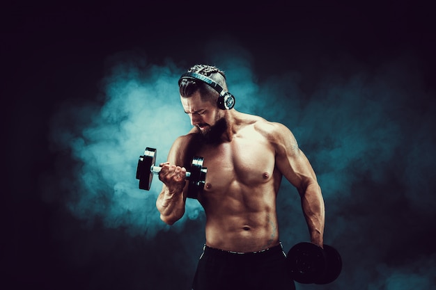 Homme formation des muscles avec des haltères en studio sur fond sombre avec de la fumée.