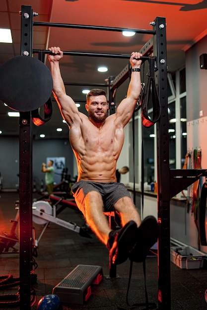 Homme fort athlétique sans chemise sur fond de gym sur la barre horizontale. Vue de face.