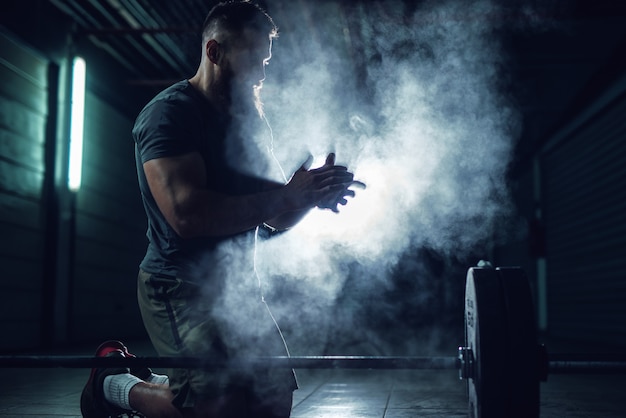 Homme fort de bodybuilder de fitness actif travailleur accroupi et applaudissant des mains avec de la poudre de craie avant l'entraînement en force avec un haltère lourd