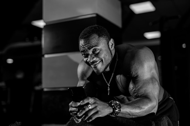 Homme fort bodybuilder tenant un smartphone et regardant la caméra tout en posant dans la salle de gym Concept d'athlète afro-américain Photo noir et blanc
