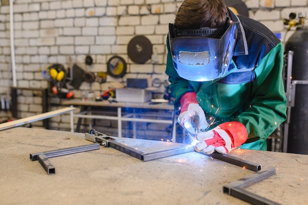 Un homme fort est un soudeur dans un masque de soudage et des cuirs de soudeurs, un produit métallique est soudé avec une machine à souder dans le garage