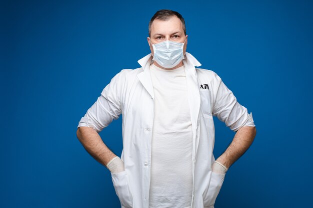 Un homme fort avec des lunettes, une blouse médicale blanche et un masque se tient les mains dans les poches