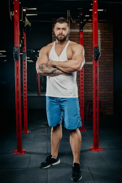 Homme fort musclé avec des tatouages et de la barbe posant dans un débardeur blanc et un short bleu dans la salle de gym.