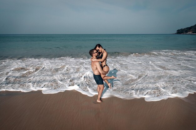 Un homme fort tient sa petite amie dans ses bras sur la belle plage au bord de la mer bleue, amour et tendresse.