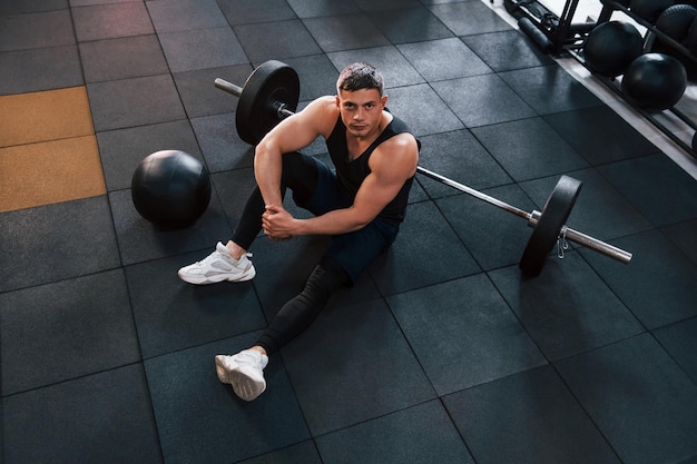 Homme fort en vêtements sportifs assis sur le sol dans la salle de sport avec équipement