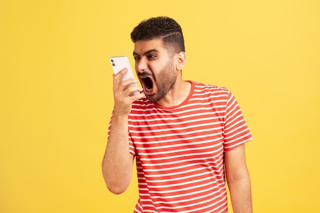 Homme fou en colère avec barbe en t-shirt rayé criant bruyamment téléphone parlant, laissant un message vocal avec une expression agressive furieuse. Tourné en studio intérieur isolé sur fond jaune