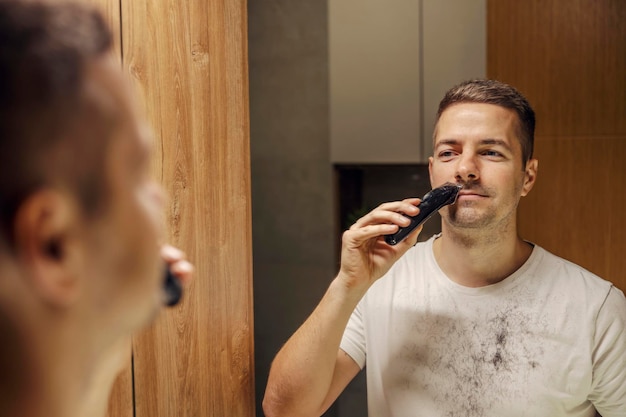 Un homme fraîchement rasé se rase la moustache en se tenant debout dans la salle de bain devant le miroir