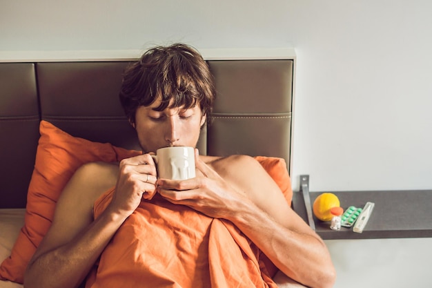 Photo l'homme a froid, allongé dans le lit et boit du thé et mesure la température avec un thermomètre