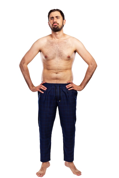 Un homme frustré se tient en pantalon avec un torse nu. Personnage antisportif. Pleine hauteur. Isolé sur fond blanc. Verticale.