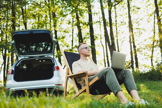 Photo un homme gai à lunettes est assis sur une chaise dans la nature et travaille en vacances en ligne sur un ordinateur portable