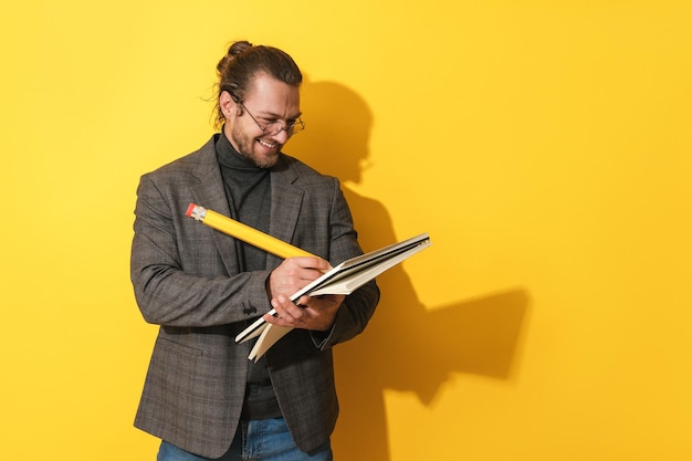 Homme gai portant des lunettes tenant un gros crayon et un cahier sur fond jaune