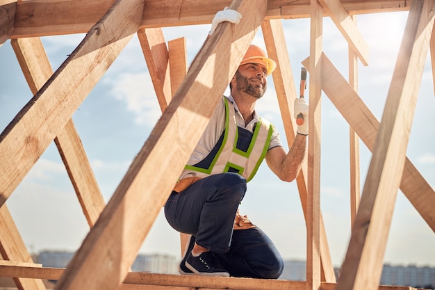 Homme gai se tenant sur un genou et frappant un clou avec un marteau tout en construisant une carcasse de toit en bois