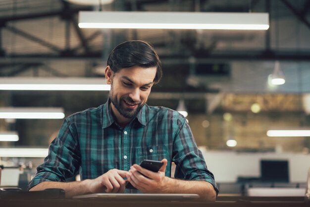 Homme gai souriant et utilisant son smartphone assis à la table