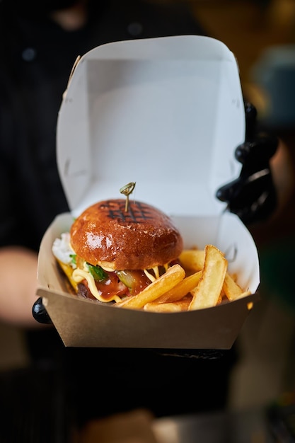 Un homme en gants noirs tient un hamburger dans ses mains Burger emballé dans une boîte en carton blanc