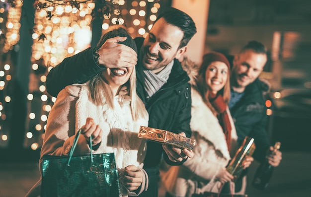 L'homme garde les yeux de sa petite amie couverts pendant qu'elle offre un cadeau. Leurs amis debout à l'extérieur avec des sacs à provisions et des boîtes. Mise au point sélective. Concentrez-vous sur le premier plan.