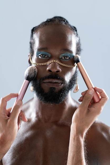 Photo homme gay barbu qui s'applique du maquillage avec des pinceaux tenus par un artiste des récoltes
