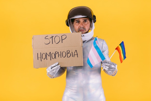 Homme gay habillé en astronaute avec un casque et un costume argenté avec des drapeaux du collectif lgtbi et trans et tenant une pancarte indiquant 39Arrêtez l'homophonie39 sur fond jaune