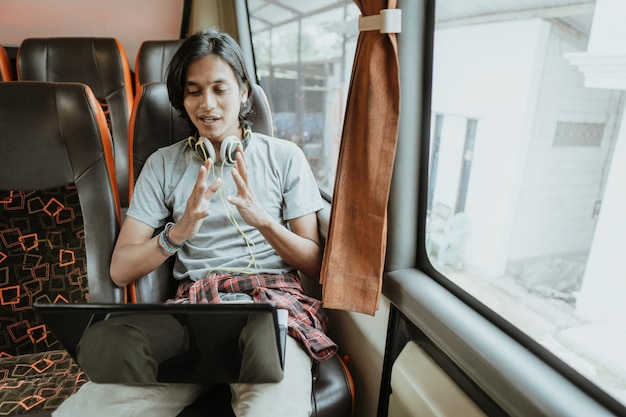 Un homme avec des gestes de la main et des écouteurs utilise un ordinateur portable tout en discutant à l'aide d'une réunion virtuelle tout en étant assis près d'une fenêtre dans un bus