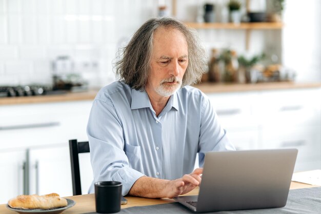 Un homme gris caucasien âgé et concentré est assis à un bureau avec un ordinateur portable dans la cuisine à la maison, travaille en ligne, vérifie et répond aux courriels, parcourt les nouvelles sur Internet, envoie des messages.