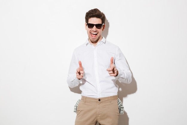 Homme guilleret en lunettes de soleil noir pointant des index sur l'appareil photo avec beaucoup d'argent billets d'un dollar qui sort des poches, isolé sur un espace blanc avec ombre