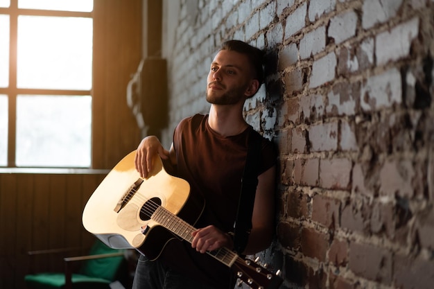 Homme avec guitare acoustique appuyé contre un mur de briques jouant de la musique en chantant des chansons profiter de la vie Beau guitariste masculin caucasien pratique jouer instrument de musique maison loft intérieur Mode de vie créatif