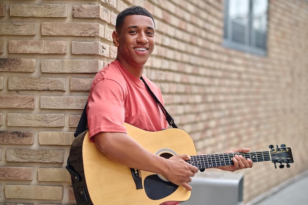 Homme avec guitare debout appuyé contre un mur de briques