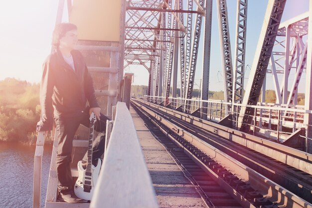 Un homme avec une guitare électrique sur le chemin de fer. Un musicien en veste de cuir avec une guitare dans la rue de la zone industrielle. Guitariste sur le pont.