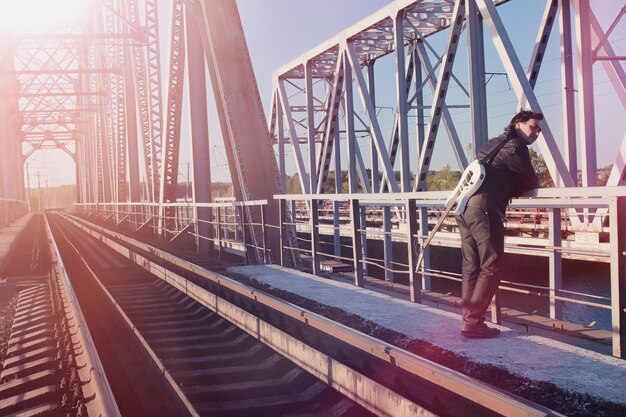 Un homme avec une guitare électrique sur le chemin de fer. Un musicien en veste de cuir avec une guitare dans la rue de la zone industrielle. Guitariste sur le pont.