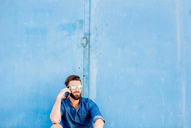 Homme habillé décontracté en chemise bleue assis avec téléphone sur le fond du mur bleu. Plan large avec espace de copie