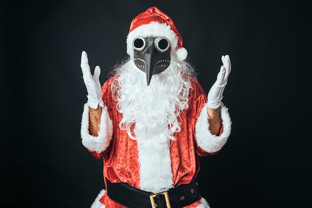 Homme habillé en Père Noël avec un masque de peste bubonique victorienne, levant les mains, sur fond noir. Concept de Noël, Père Noël, cadeaux, célébration.