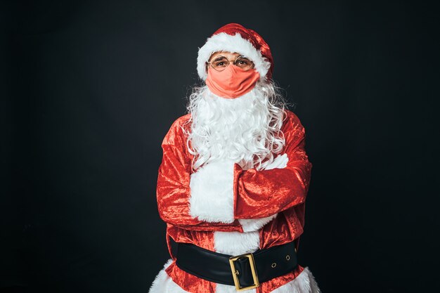 Homme habillé en Père Noël avec un masque rouge et les bras croisés, sur fond noir. Concept de Noël, Père Noël, cadeaux, célébration.