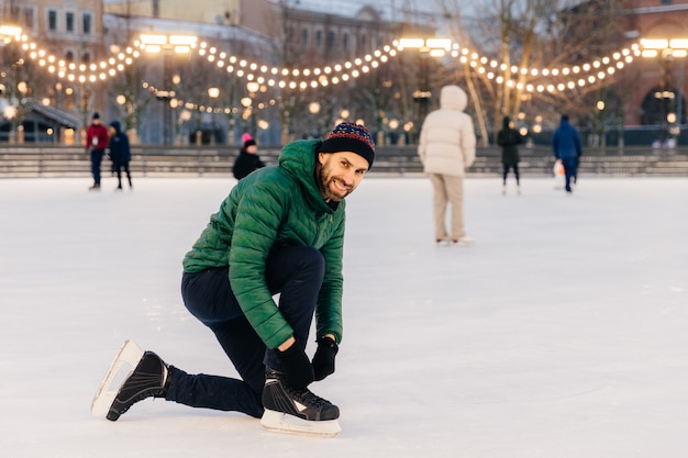 Homme en habit vert, lace des patins, regarde avec bonheur dans l'appareil photo, se dresse sur un anneau magnifique