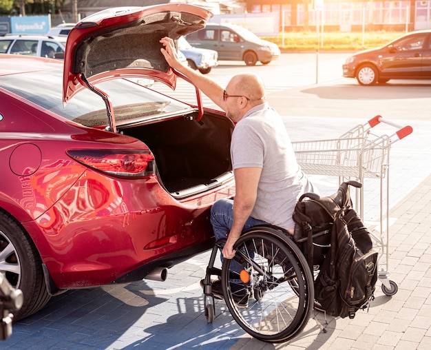 L'homme handicapé adulte dans un fauteuil roulant met des achats dans le coffre d'une voiture dans un parking de supermarché