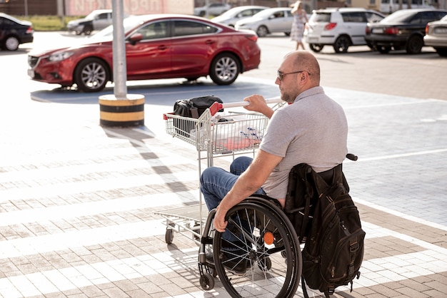 L'homme handicapé adulte dans un fauteuil roulant pousse un chariot vers une voiture dans un parking de supermarché