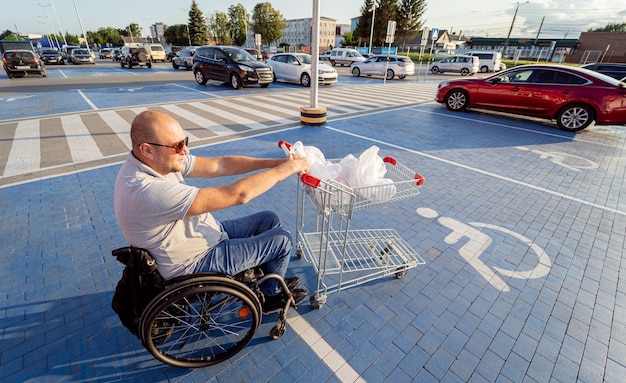 L'homme handicapé adulte dans un fauteuil roulant pousse un chariot vers une voiture dans un parking de supermarché