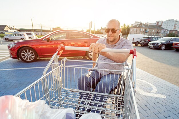 Photo homme handicapé en fauteuil roulant poussant le chariot devant lui au parking du supermarché