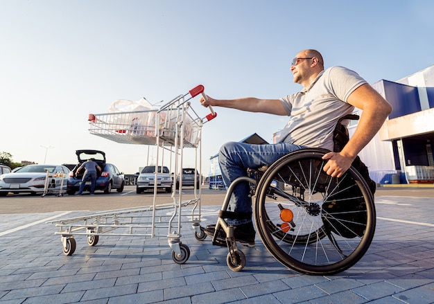 Homme handicapé en fauteuil roulant poussant le chariot devant lui au parking du supermarché