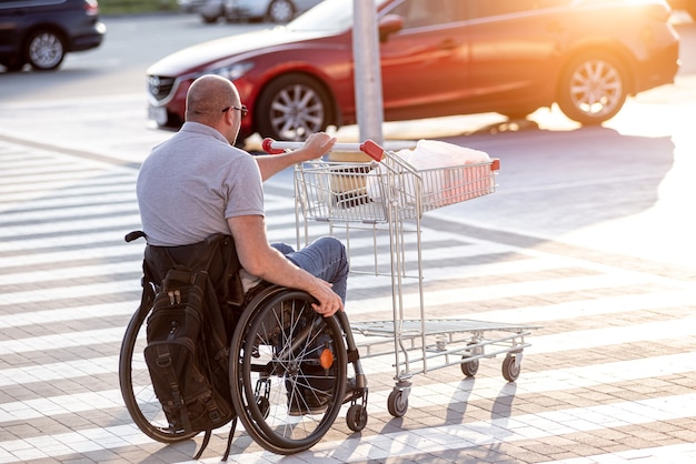 Homme handicapé en fauteuil roulant poussant le chariot devant lui au parking du supermarché