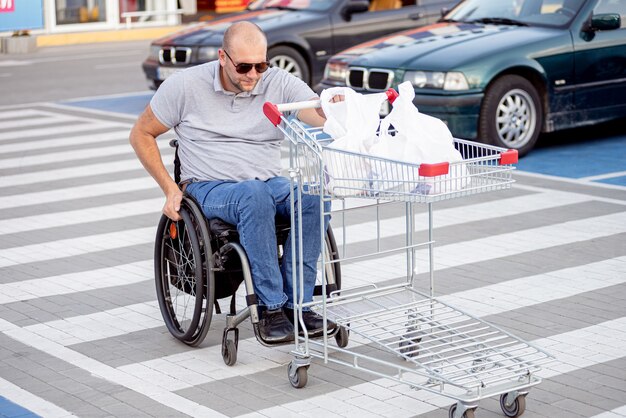 Homme handicapé en fauteuil roulant poussant le chariot devant lui au parking du supermarché