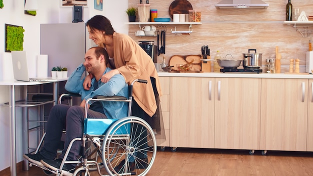 Homme handicapé souriant en fauteuil roulant et sa femme lors d'un appel vidéo sur ordinateur portable dans la cuisine. Homme d'entreprise avec paralysie handicap handicap handicapé difficultés de travail après accident ayant stagiaire