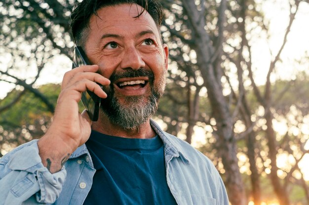 Un homme heureux adulte souriant et parlant au téléphone en plein air dans le parc avec la lumière du coucher du soleil et des arbres en arrière-plan Des hommes en denim décontractés utilisant leur téléphone portable pour appeler des amis Portrait joyeux de la personne