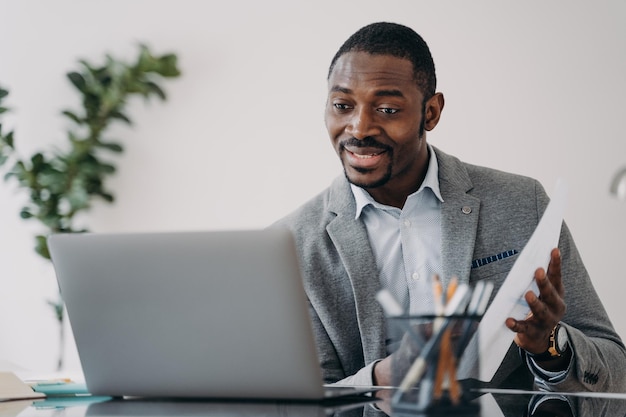 Un homme heureux a un appel vidéo sur un ordinateur portable et pointe vers des documents de données Exécutif africain à distance