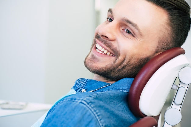 Un homme heureux aux yeux noisette et aux cheveux brun foncé est assis dans un fauteuil dentaire et sourit directement dans la caméra parce qu'il est satisfait du travail d'un dentiste