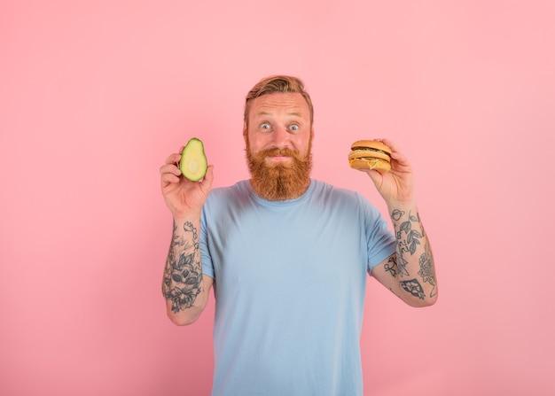 L'homme heureux avec la barbe et les tatouages est indécis s'il doit manger un avocat ou un hamburger