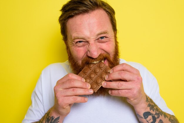 L'homme heureux avec la barbe et les tatouages mange du chocolat