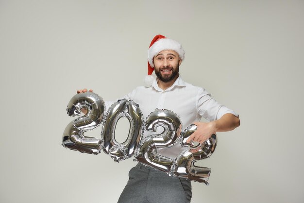 Un homme heureux dans un bonnet de noel en velours rouge montre des ballons argentés en forme de 2022. Un gars souriant avec une barbe lors d'une fête du nouvel an.